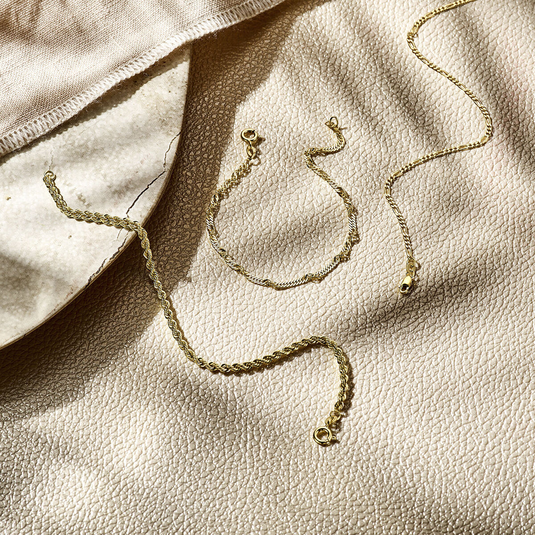 Drie delicate 14K geelgouden sieraden, waaronder de Van der Kooij Collectie armband figaro 1,8 mm, liggen op een beige oppervlak met zachte schaduwen en een stukje lichte stof zichtbaar in de hoek.