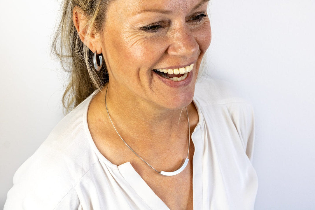 Een lachende vrouw met lang haar draagt een witte blouse en zilveren sieraden, waaronder de uniek ontworpen Collier Bogen C74-ketting van CLIC Creations met een magnetische sluiting en oorringen, tegen een effen witte achtergrond.