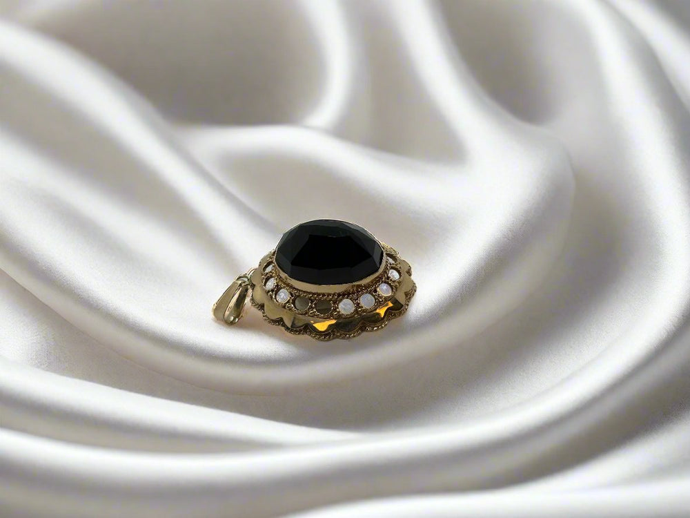 Op gladde witte zijde ligt een sierlijke 14 karaat gouden ring van Juwelier van der Kooij, met een grote zwarte edelsteen omringd door parelachtige accenten, wat doet denken aan een vintage collectie.