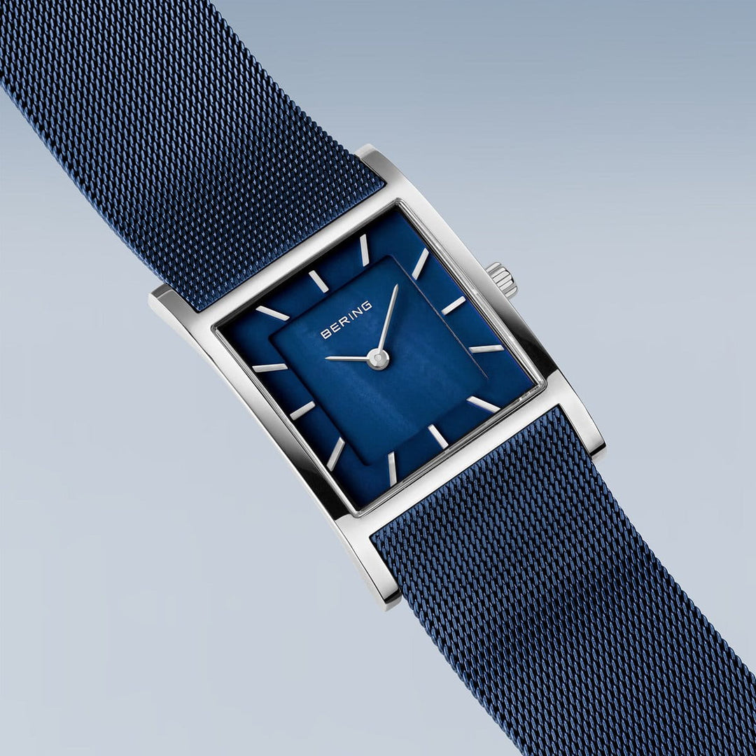 Het Bering dameshorloge 10426-307-S heeft een rechthoekig ontwerp met een blauwe wijzerplaat en meshband. Het bevat zilveren uurmarkeringen, wijzers en merknaam, afgezet tegen een lichtblauwgrijze achtergrond met kleurverloop. De zilveren kast wordt beschermd door duurzaam saffierglas en wordt aangestuurd door quartz uurwerk.