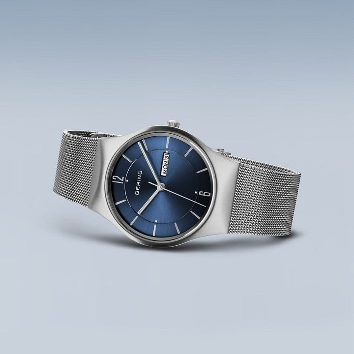 Het Bering 11938-003DD Herenhorloge is een strak zilveren horloge met een blauwe wijzerplaat, Saffierglas, zilveren uurmarkeringen en wijzers, dag- en datumaanduiding en een meshband. Dit alles steekt elegant af tegen een lichtblauwe achtergrond voor een minimalistisch design.