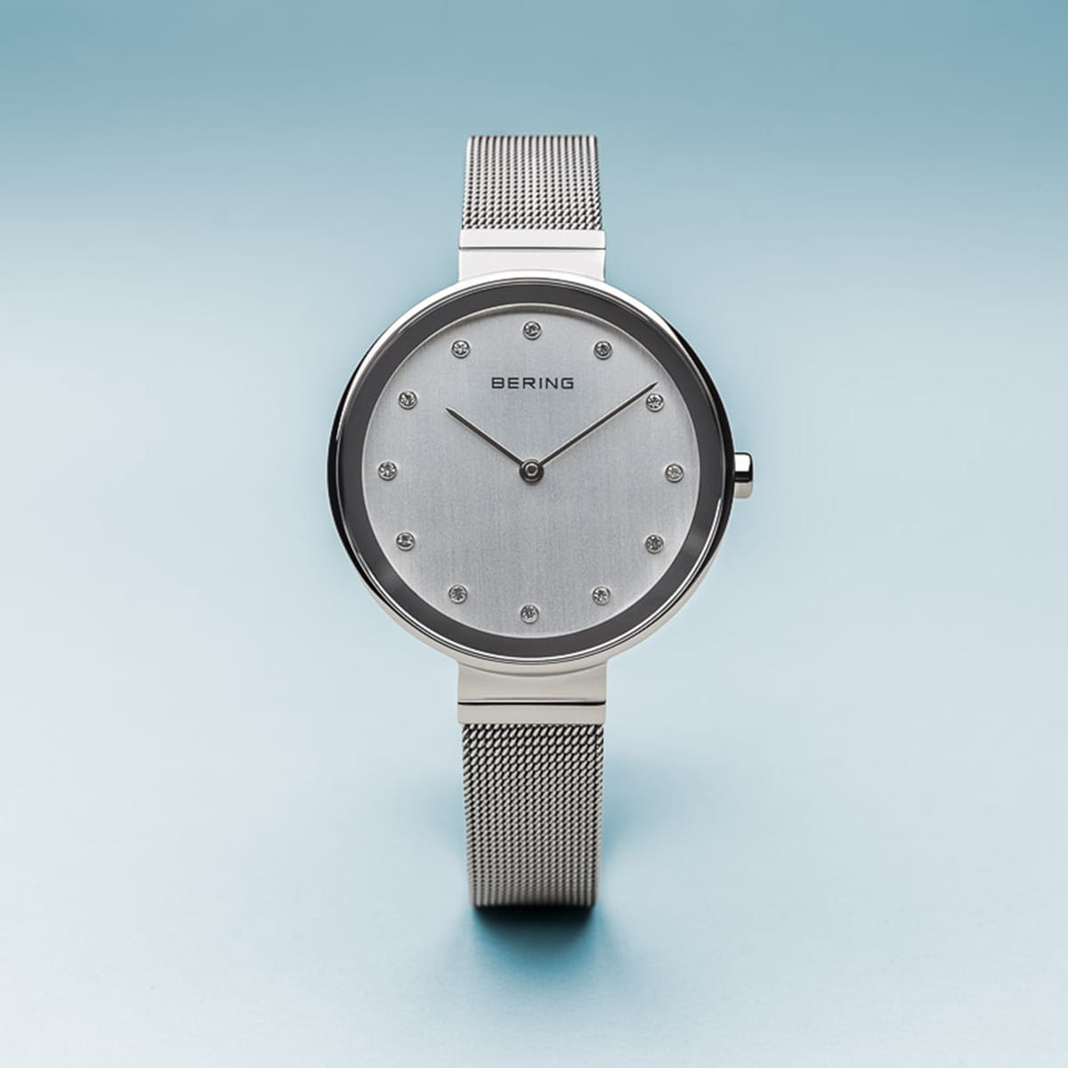 Het Bering 12034-000 horloge heeft een minimalistisch zilver design met een ronde wijzerplaat, stud uurmarkeringen en slanke wijzers. Het heeft Saffierglas en Quartz-uurwerk, een mesh band tegen een gradiënt blauwe achtergrond en de merknaam Bering wordt weergegeven.