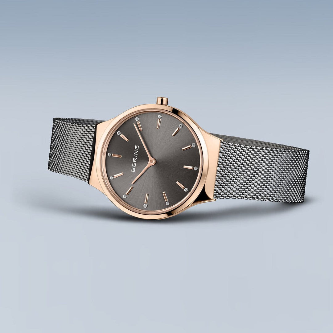 Het Bering Dameshorloge (Product 12131-369-GWP) heeft een roségouden lunette en een grijze wijzerplaat met minimalistische markeringen. Het heeft een fijne zilveren Milanees mesh band. Dit quartz-uurwerk is elegant geplaatst tegen een vloeiende blauwe achtergrond.