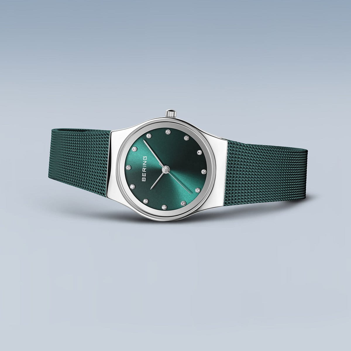 Het Bering Dameshorloge, model 12927-808, heeft een groene wijzerplaat met puntmarkeringen en het BERING-logo. De zilveren kast is gecombineerd met een getextureerd Milanees bandje, allemaal op een grijze achtergrond, wat de tijdloze Quartz-uurwerkprecisie belichaamt.