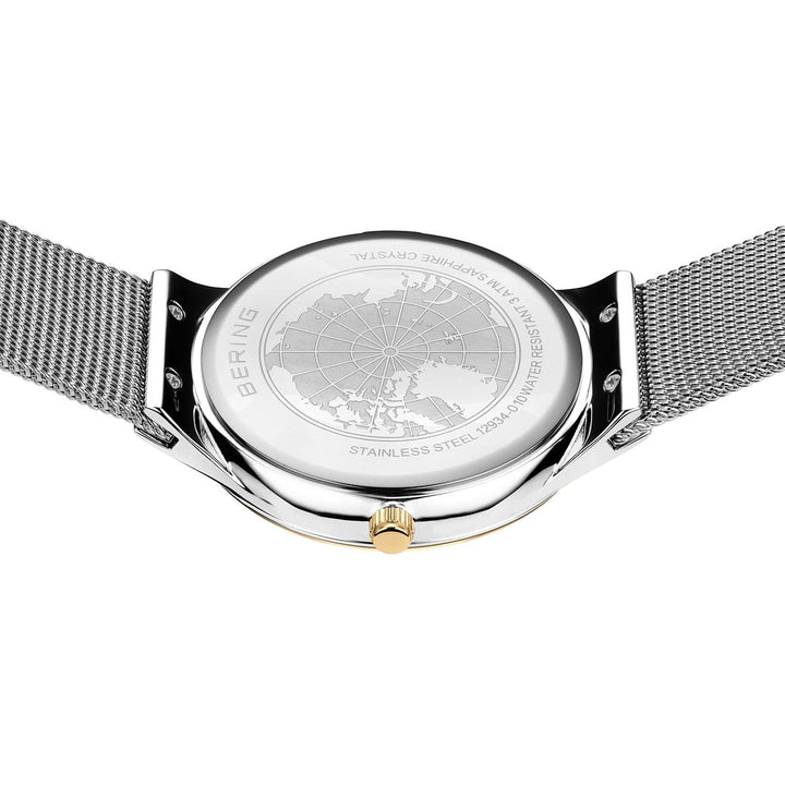 De afbeelding toont de achterkant van een Bering 12934-010 Dameshorloge met een tijdloos design, met een roestvrijstalen kast gegraveerd met een wereldbol en tekst over materialen en waterbestendigheid. Een metalen meshband voegt elegantie toe.