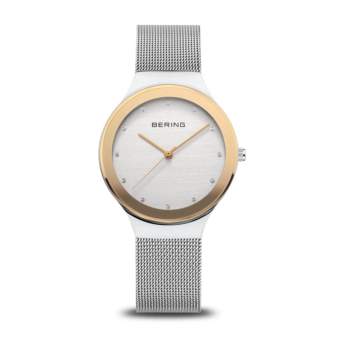 Berings 12934-010 Dameshorloge toont een tijdloos design met een zilveren meshband, ronde wijzerplaat, roestvrijstalen kast, gouden lunette, minimalistische uurmarkeringen en twee wijzers. Bering is elegant gepositioneerd onder de 12-uursmarkering.