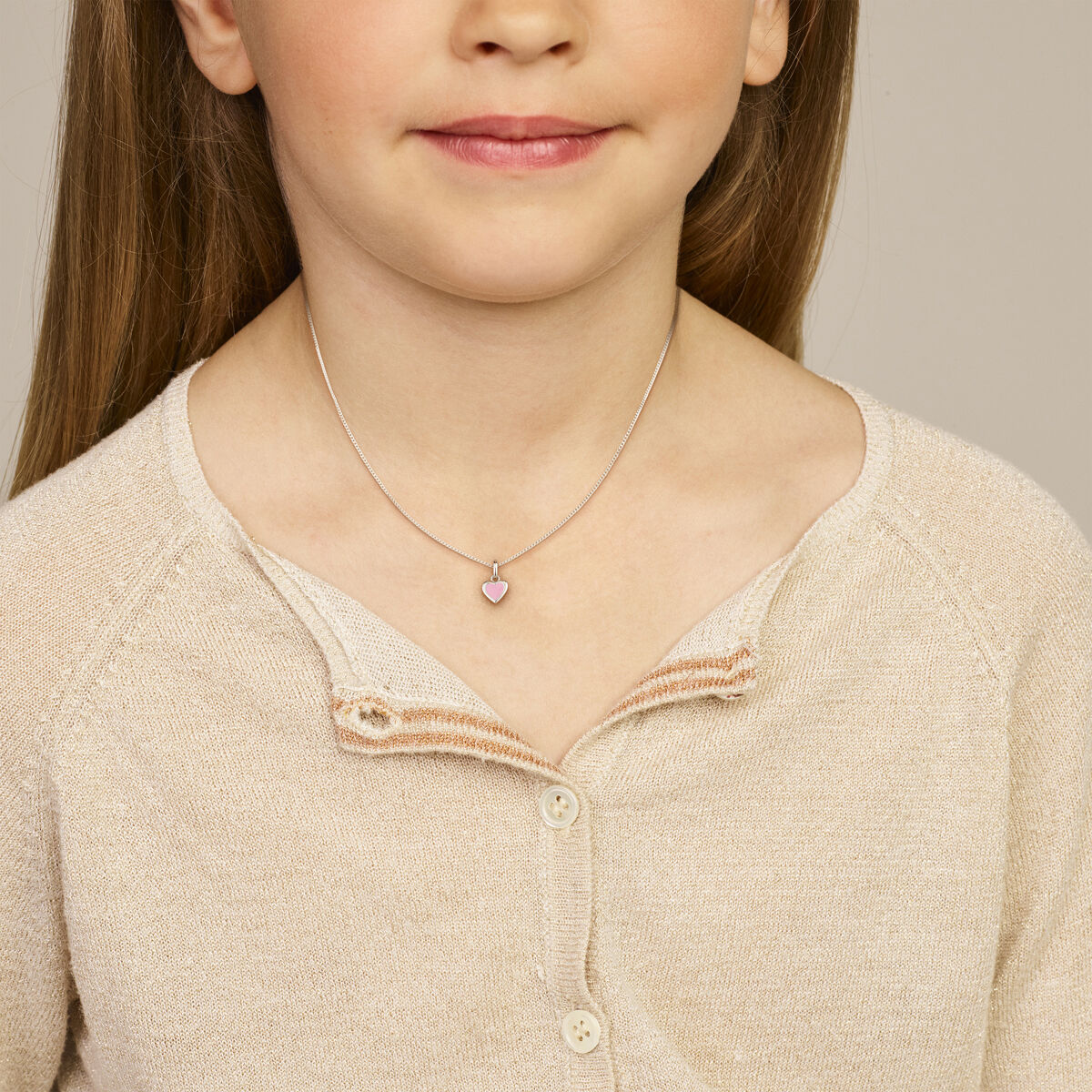 Een jong meisje draagt een crèmekleurige trui en de Van der Kooij Collectie hanger hart zilver gerhodineerd, een delicate zilveren harthanger – zichtbaar vanaf de schouders, zachtjes glimlachend. Perfect sieraad voor kinderen.