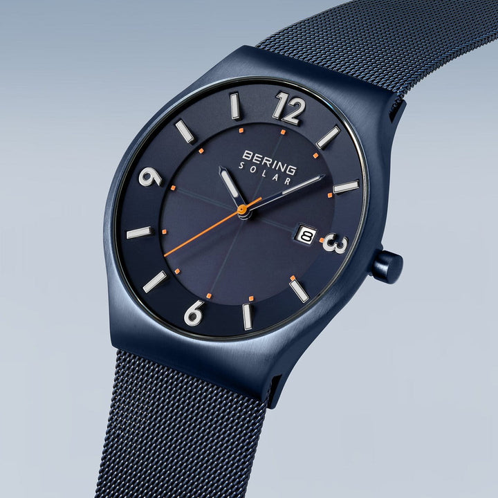 Het Bering 14440-393 horloge heeft een blauwe metalen mesh band en wijzerplaat met Arabische cijfers op 3, 6, 9 en 12 uur. Het heeft oranje uurmarkeringen en secondewijzer, een Solar logo naast de merknaam op de wijzerplaat, alles beschermd door duurzaam saffierglas.