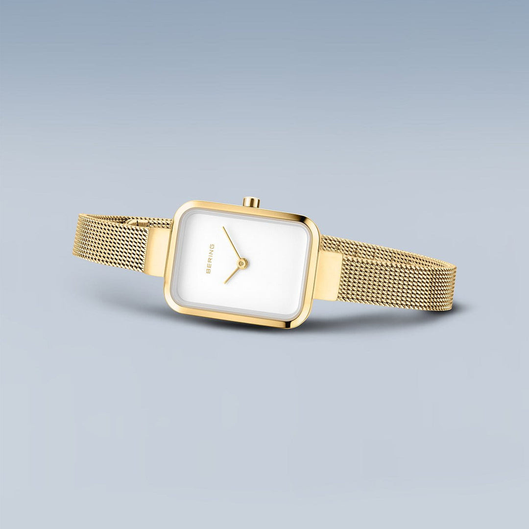 De Bering 14520-334 dameshorloge heeft een minimalistisch goudkleurig ontwerp met een vierkante witte wijzerplaat, roestvrijstalen kast, goudkleurige mesh band en eenvoudige wijzers op een lichtgrijsblauwe achtergrond.