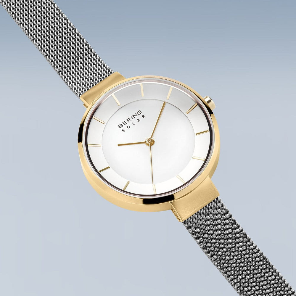 Het Bering dameshorloge 14631-024 heeft een goudkleurige kast met een minimalistische witte wijzerplaat, gouden uurmarkeringen en quartz-uurwerk. Het heeft een zilveren meshband en is elegant geplaatst tegen een zachtblauwe gradiëntachtergrond.