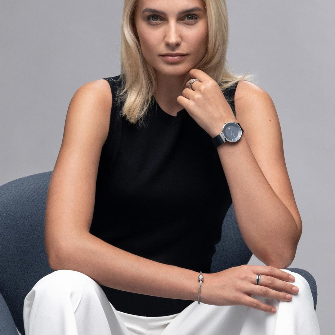 Een persoon met lang blond haar zit op een stoel, draagt een zwart mouwloos topje en een witte broek. Ze laten hun kin op hun hand rusten en tonen het gestroomlijnde Bering 14639-307 horloge met saffierglas en zilveren sieraden tegen een zachtgrijze achtergrond.