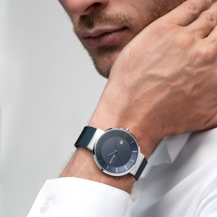 Een man in een wit overhemd, in een minimalistische setting, rust zijn kin op zijn hand, en toont het Bering horloge 14639-307 met een donkere wijzerplaat en mesh band. Het horloge is elegant voorzien van Saffierglas en solar-uurwerk technologie.