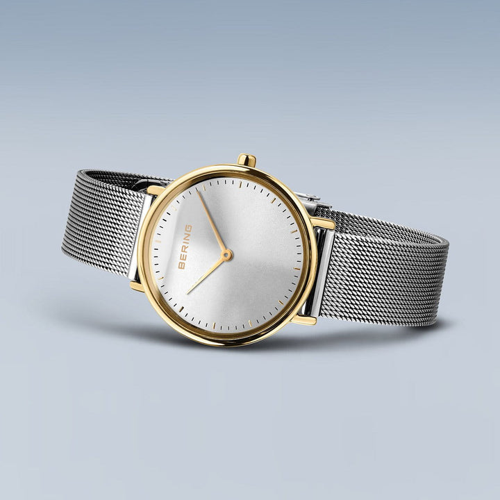De Bering 15729-010, een minimalistisch polshorloge, heeft een witte wijzerplaat zonder cijfers, een goudkleurige kast, slanke gouden wijzers met Quartz-technologie voor precisie en een zilveren meshband op een rustgevende blauwe achtergrond.