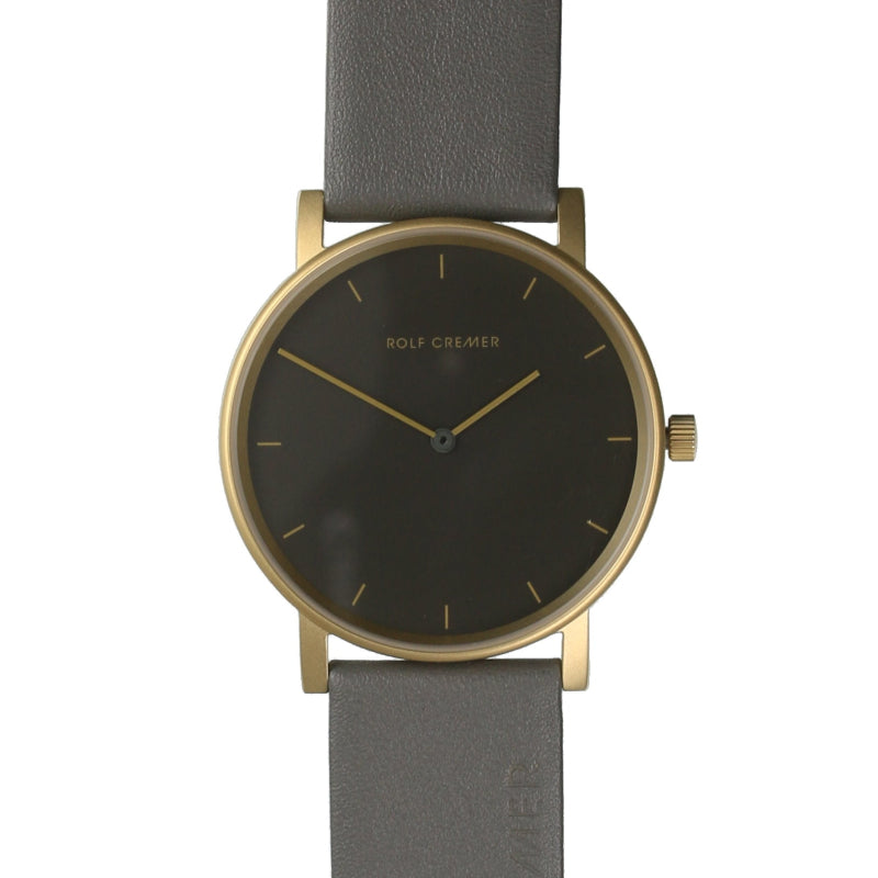 Montre Rolf Cremer avec boîtier doré, cadran noir et bracelet en cuir gris.