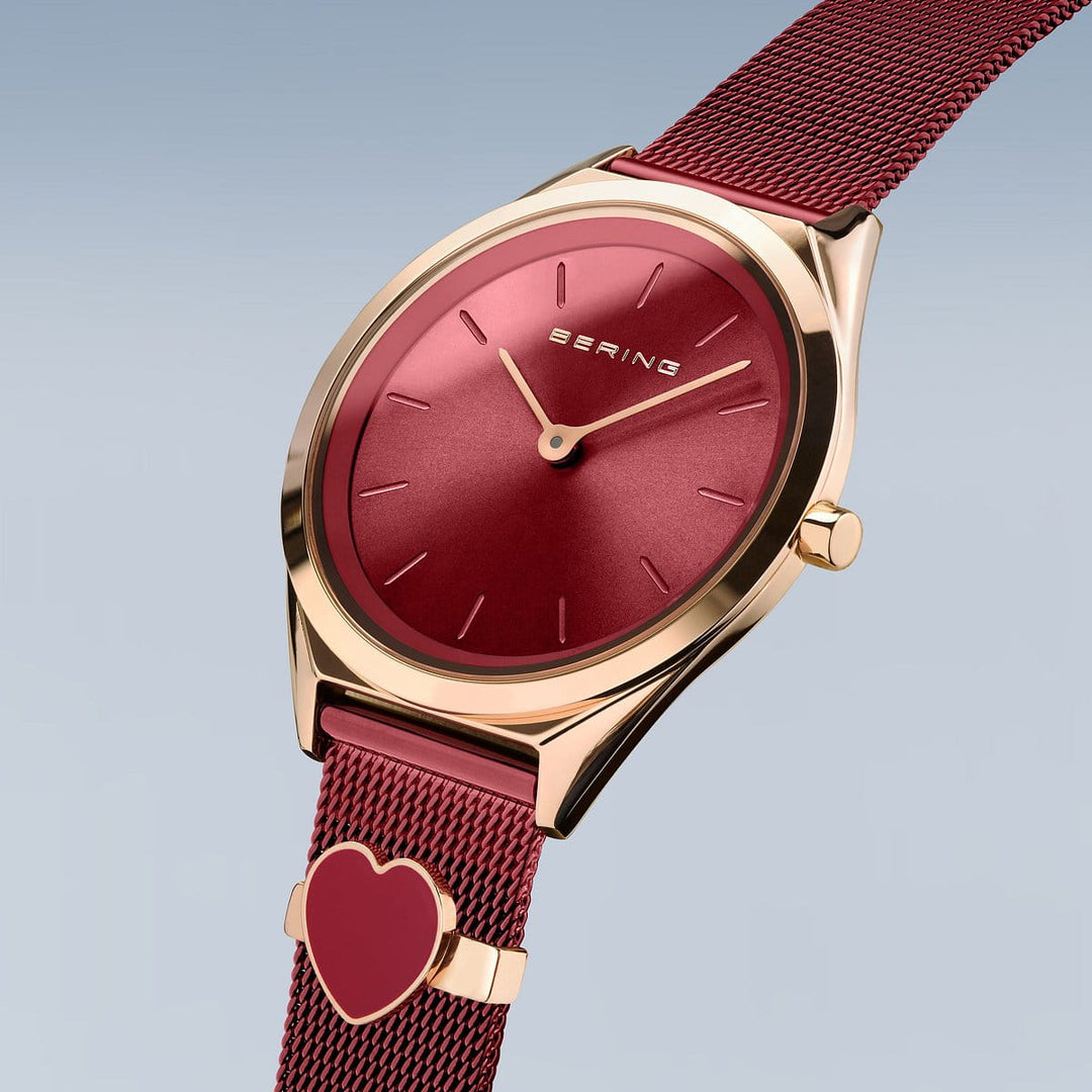 Het Bering 17031-363-GWP dameshorloge heeft een minimalistische goud-rode wijzerplaat met logo, die op een rood Milanees bandje zit. De elegante rode mesh band bevat een hartvormige bedel, afgezet tegen een lichtblauwe naar grijze gradiënt achtergrond.