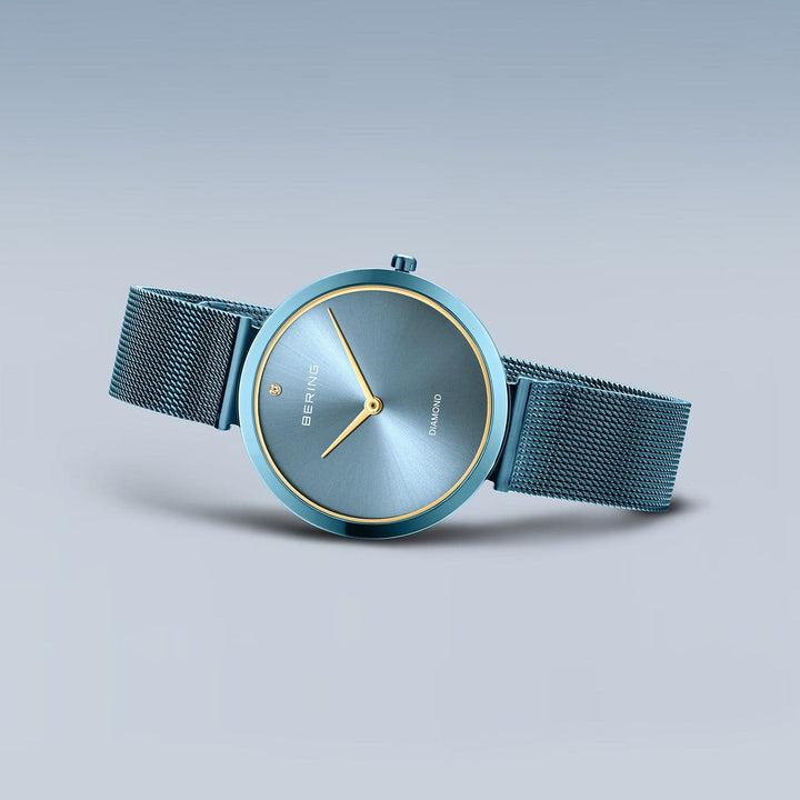 De Bering dameshorloge 18132-Charity1 heeft een blauwe mesh band, minimalistische blauwe wijzerplaat, gouden markers en wijzers en duurzaam saffierglas. Het Bering-logo verschijnt op de wijzerplaat op een lichtblauwe achtergrond met kleurverloop.