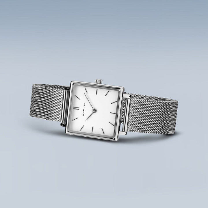 Het Bering dameshorloge 18226-004 toont een minimalistisch vierkant zilveren ontwerp met een quartz uurwerk, duurzaam saffierglas, dunne uurmarkeringen en een subtiele kroon aan de zijkant. Elegant geplaatst op een lichtblauwe achtergrond, heeft het een strakke meshband en een witte wijzerplaat.