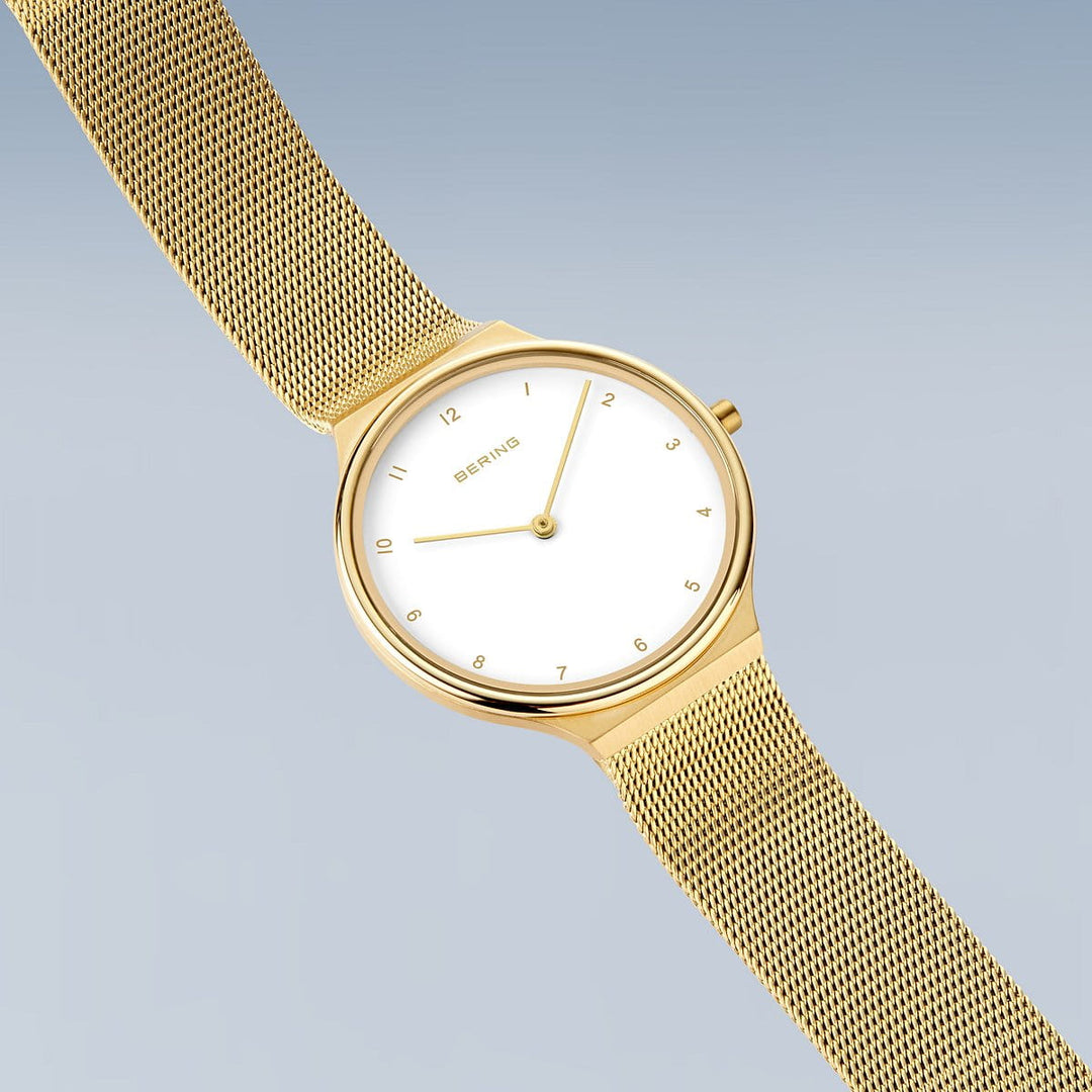 Het Bering dameshorloge goud - 18434-334 heeft een minimalistisch design met een witte wijzerplaat, gouden uurmarkeringen en een meshband, tegen een zachte blauwgrijze achtergrond.