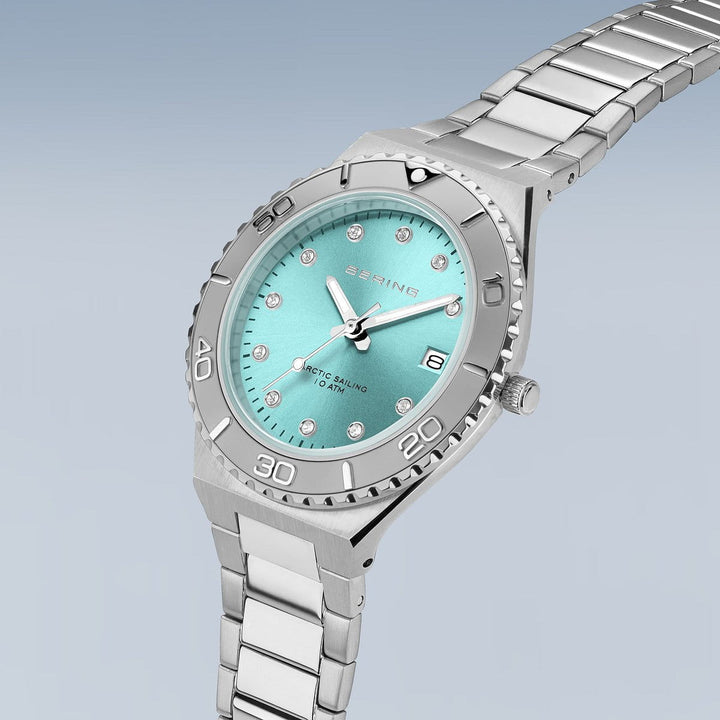 Het Bering dameshorloge 18936-707 heeft een turquoise wijzerplaat met diamantachtige markeringen, een lunette met minutenmarkeringen en een zilveren metalen band. Het biedt 10 ATM waterbestendigheid, toont BERING en ARCTIC SAILING op de wijzerplaat en is afgewerkt met duurzaam saffierglas op een lichtblauwe achtergrond met kleurverloop.