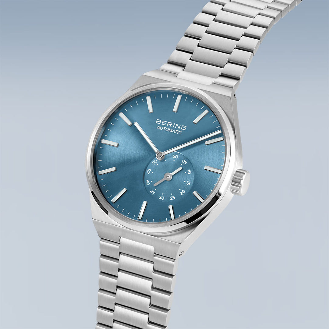 Het Bering herenhorloge 19441-Charity heeft een strakke zilveren metalen schakelband en een minimalistisch ontwerp met een blauwe wijzerplaat, saffierglas, zilveren uurmarkeringen, wijzers en een kleine secondewijzer. De wijzerplaat toont Bering en Automatic.