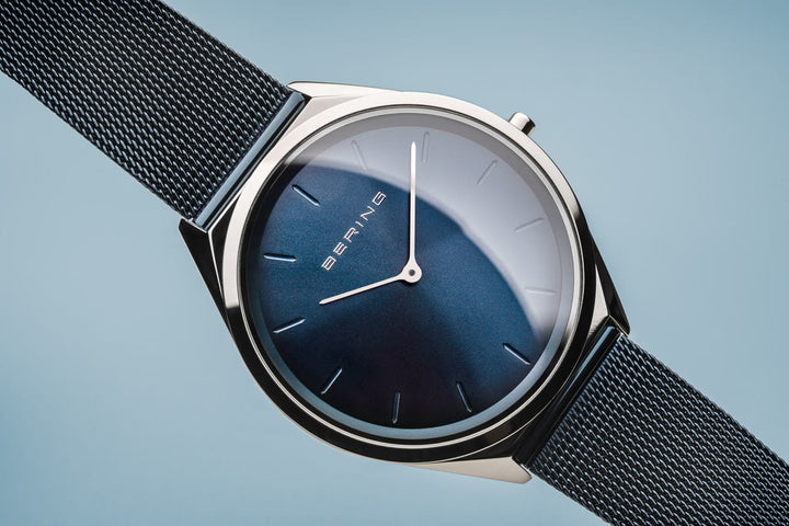 Het Bering unisex horloge (17039-307) heeft een strak minimalistisch design, met een donkerblauwe wijzerplaat met dunne zilveren uurmarkeringen onder saffierglas. Het heeft een quartz uurwerk in een slanke zilveren kast en een fijne zwarte mesh band tegen een lichtblauwe achtergrond.
