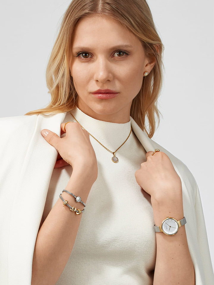 Een langharige blondine draagt een witte jas over een licht topje en draagt daarbij een gouden ketting, armbanden en oorstekers. Daarnaast draagt ze het Bering dames horloge witte dialekt - 14631-024 met quartz-uurwerk en saffierglas. Ze poseert tegen een effen achtergrond.