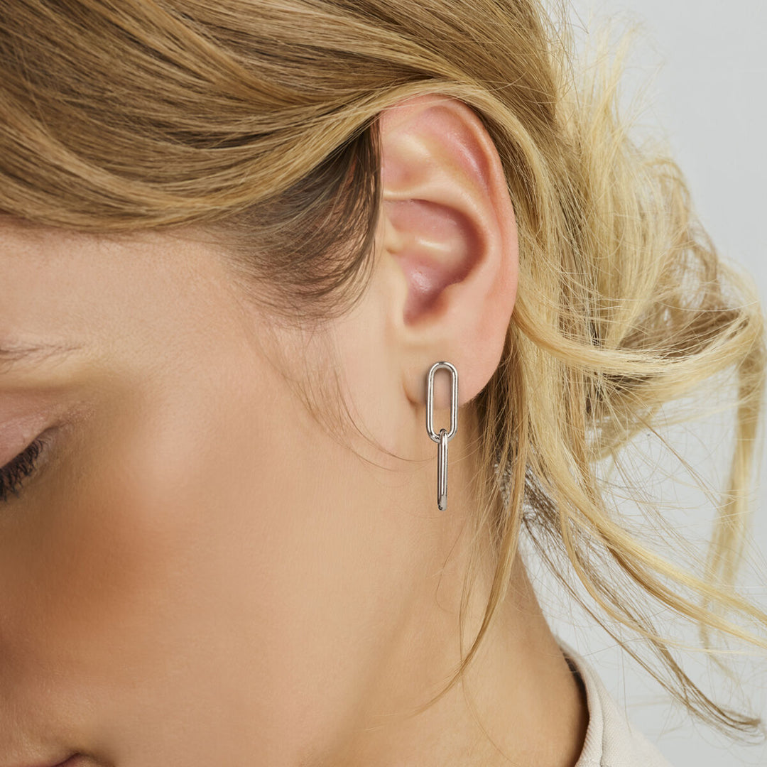 Een close-up van het oor van een vrouw met Van der Kooij Collectie oorhangers paperclip zilver gerhodineerd, voorzien van geometrische druppelvormen met rechthoekige en verticale streepjes. Haar losjes gestylde blonde haar bedekt gedeeltelijk deze elegante dames oorbellen.