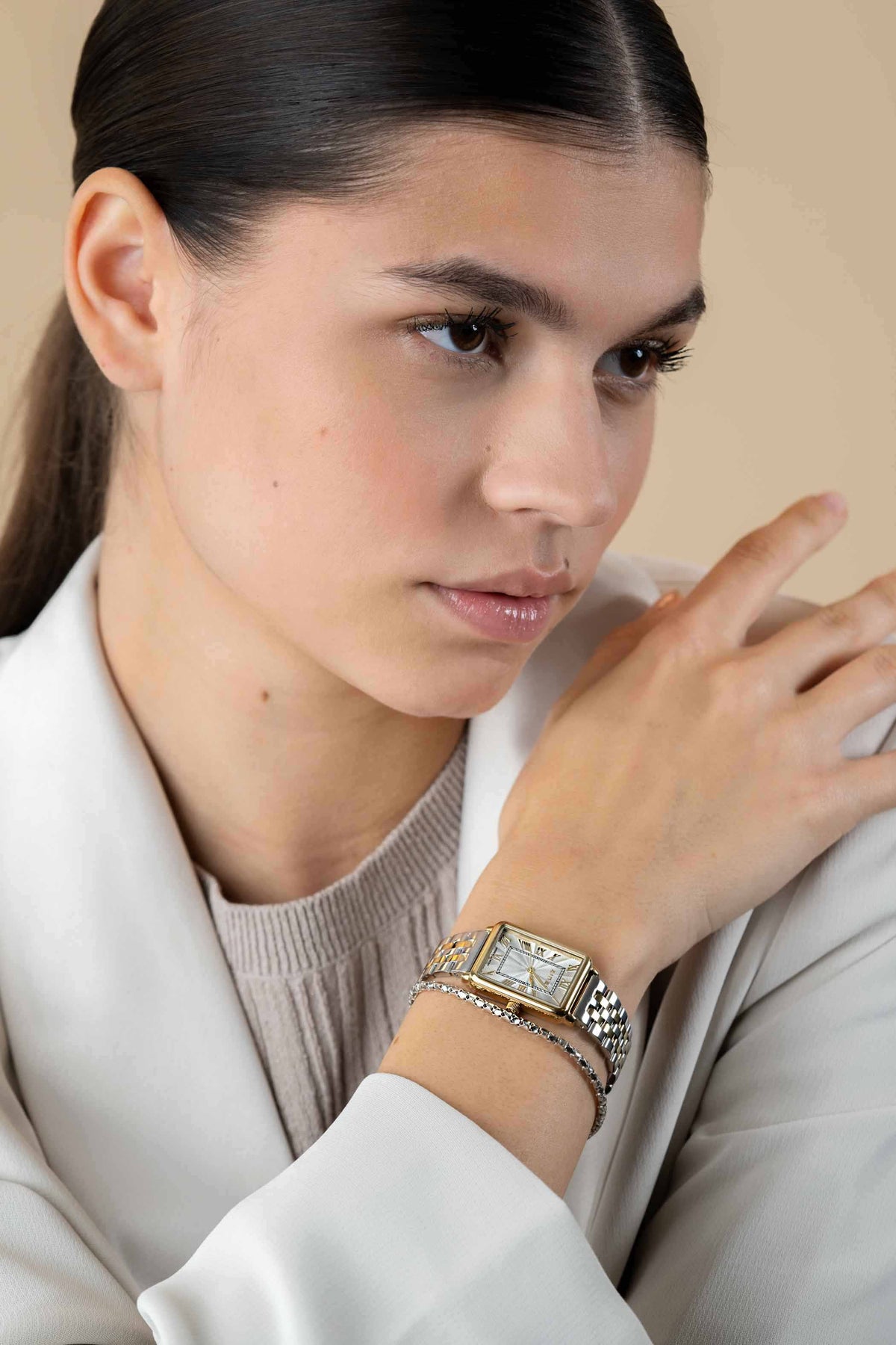 Een vrouw met steil donker haar draagt een witte blazer en een Zinzi Geelgoudkleurig Horloge Staal Ziw1907. Ze poseert met haar hand bij haar kin en kijkt nadenkend opzij.