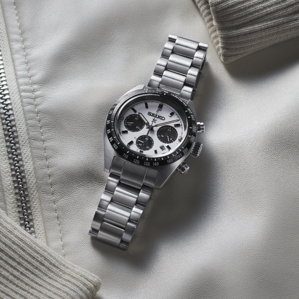 Een Seiko Prospex herenhorloge SSC813P1 met roestvrij staal, solar uurwerk en saffierglas zit elegant op een lichtgekleurde jas met rits, met een opvallende wit-zwarte wijzerplaat met drie subwijzerplaten en een strakke metalen band.
