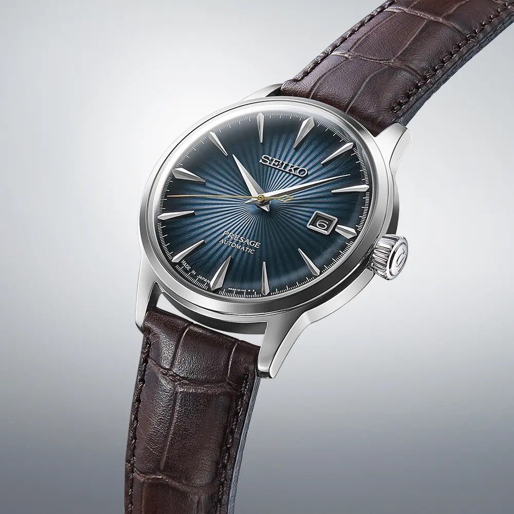 Close-up van een Seiko Presage SRPK15J1 met een metallic blauwe sunburst wijzerplaat, zilveren uurmarkeringen en datumvenster. Dit automatische horloge heeft een bruine leren band met krokodillenpatroon en zilveren wijzers met Seiko elegant gepositioneerd onder 12 uur.
