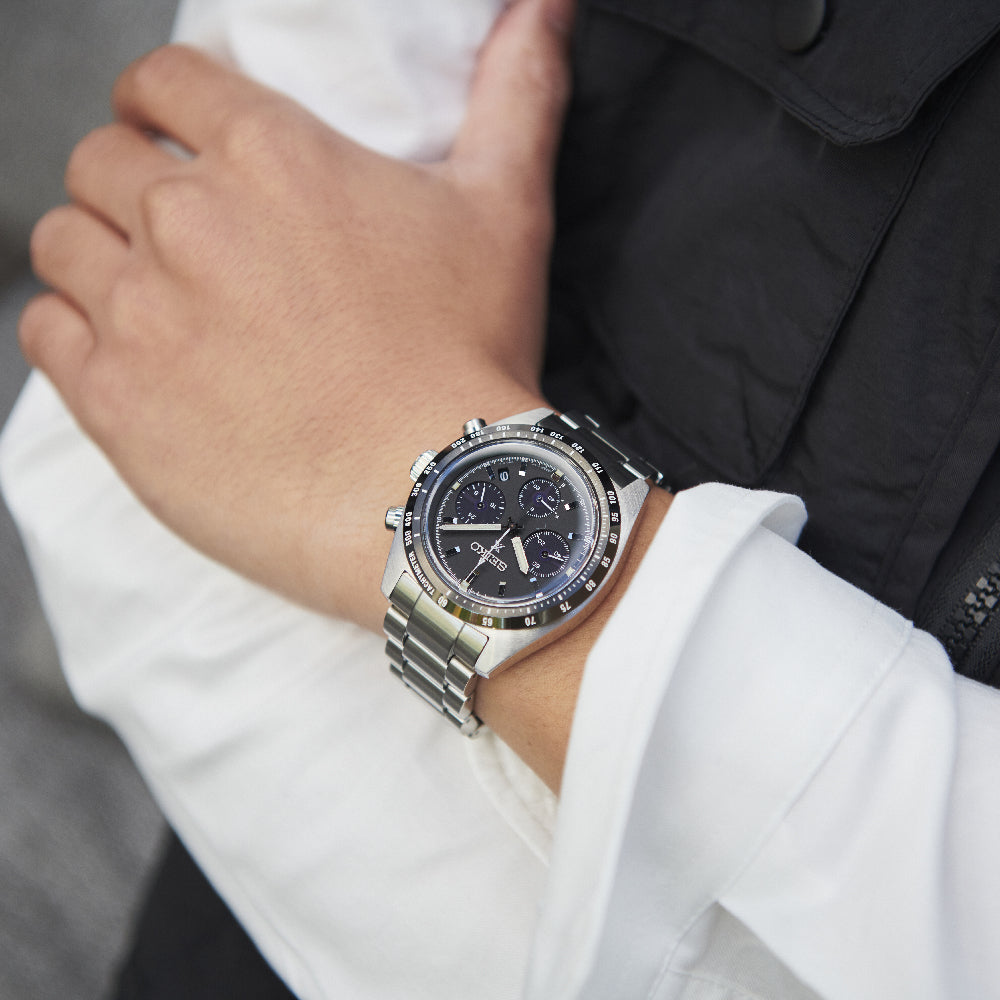 Een close-up toont een persoon die het Seiko Prospex heren horloge SSC819P1 draagt met een zwarte wijzerplaat en zilveren wijzers. De roestvrijstalen horlogekast zit op de linker pols over een witte mouw, terwijl ze de band met hun rechterhand verstellen.