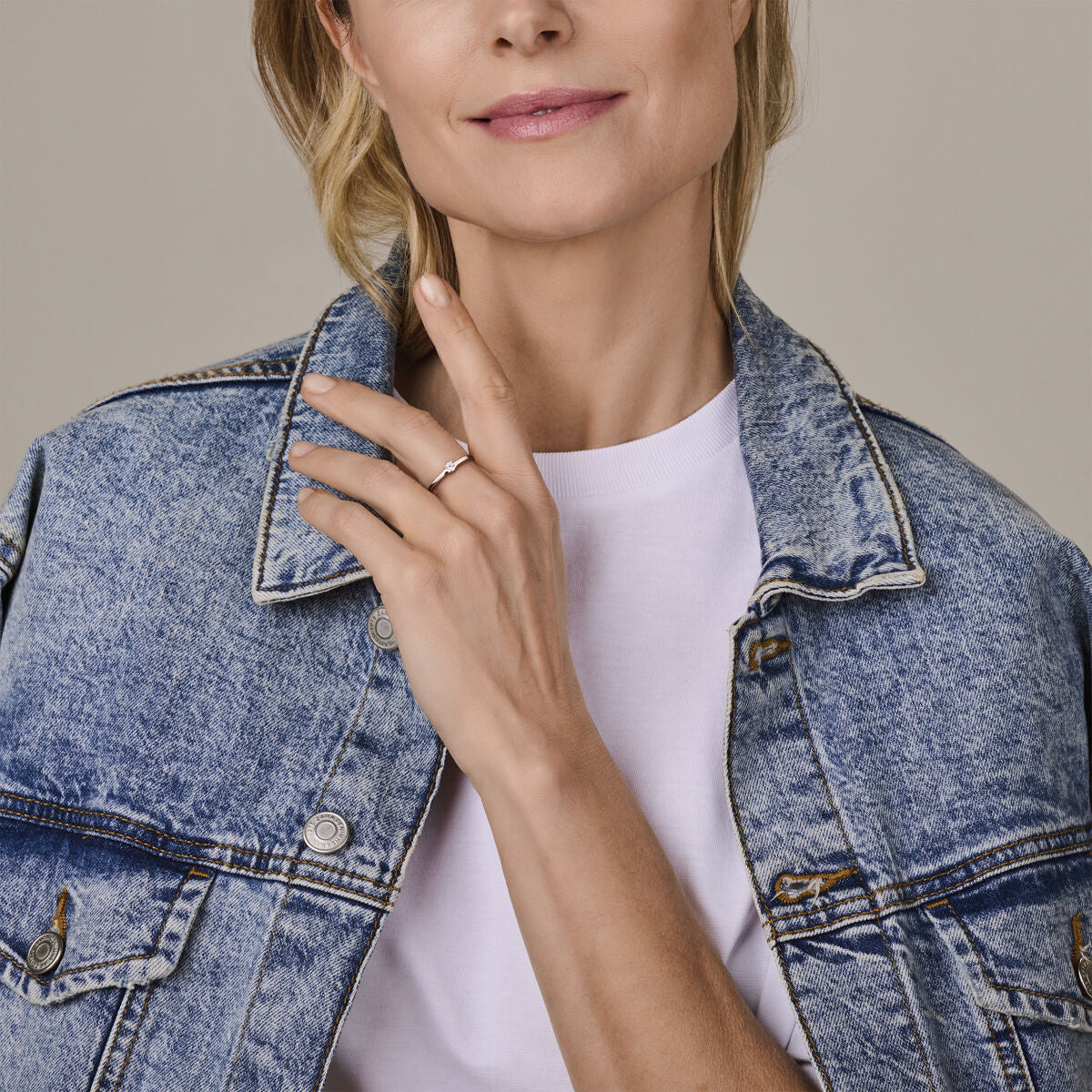 Een vrouw in een licht spijkerjasje en wit T-shirt raakt zachtjes haar gezicht aan en toont de Van der Kooij Collectie ring met geboortesteen cz april zilver gerhodineerd. Ze glimlacht subtiel, haar lichtbruine haar naar achteren getrokken.