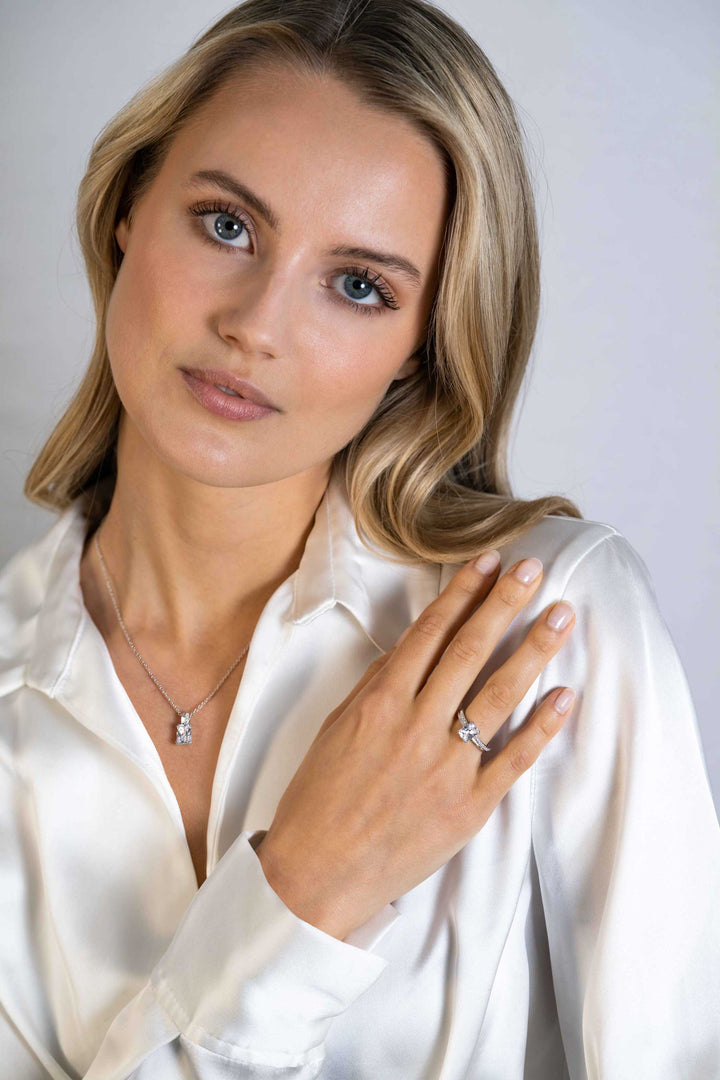 Een vrouw met lang blond haar in een witte satijnen blouse poseert met haar hand op haar borst, draagt de Zinzi Ring Zilver Zir2392 van Zinzi en een diamanten halsketting. Ze kijkt kalm en recht in de camera, tegen een lichte achtergrond.