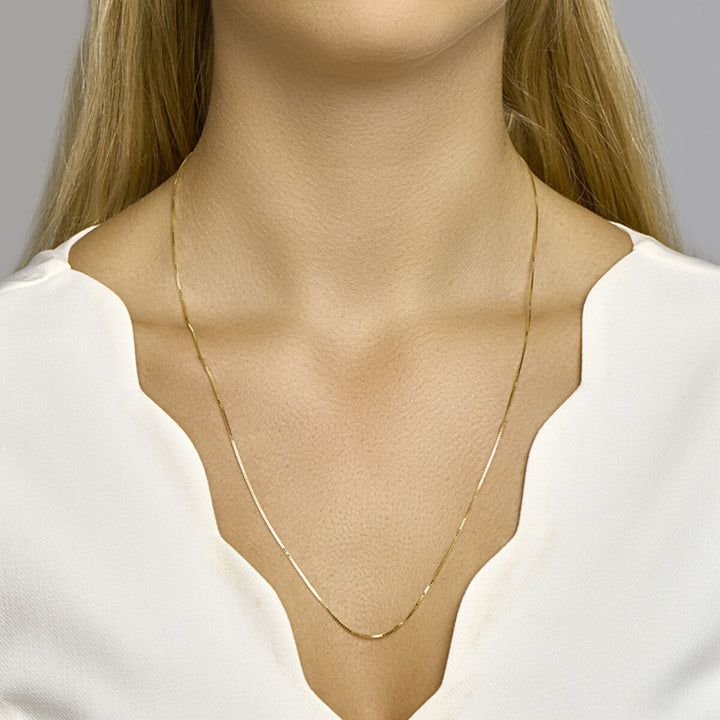 Een vrouw draagt het Van der Kooij Collectie collier venetiaans 0,8 mm 14K geelgoud van de schouders tot de borst in een wit topje met golvende halslijn tegen een neutrale achtergrond.