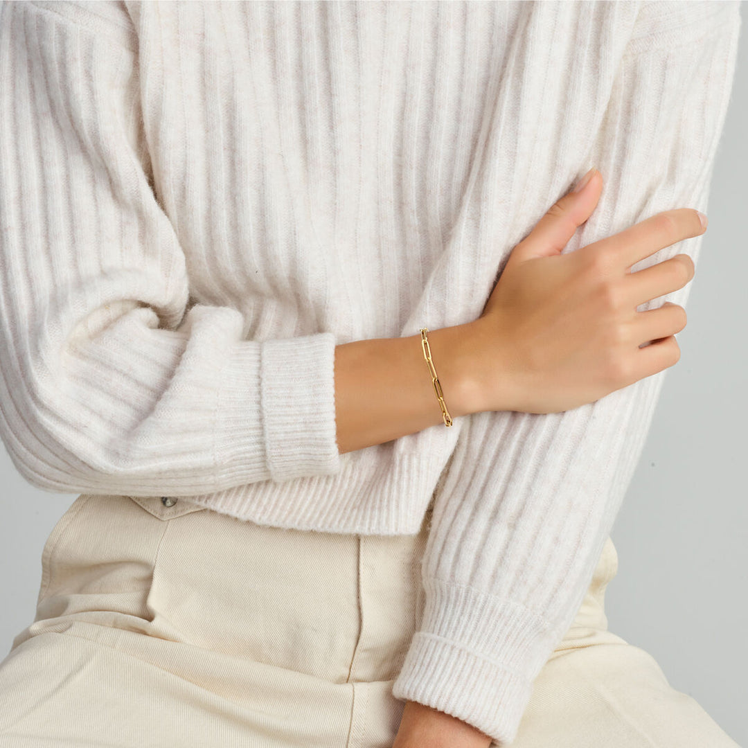 Een persoon in een geribde crèmekleurige trui met lange mouwen en een lichtbeige broek, zittend met één arm gekruist, showt een Van der Kooij Collectie Gouden armband dames paperclip vierkante buis met groot springslot 14K om zijn pols.