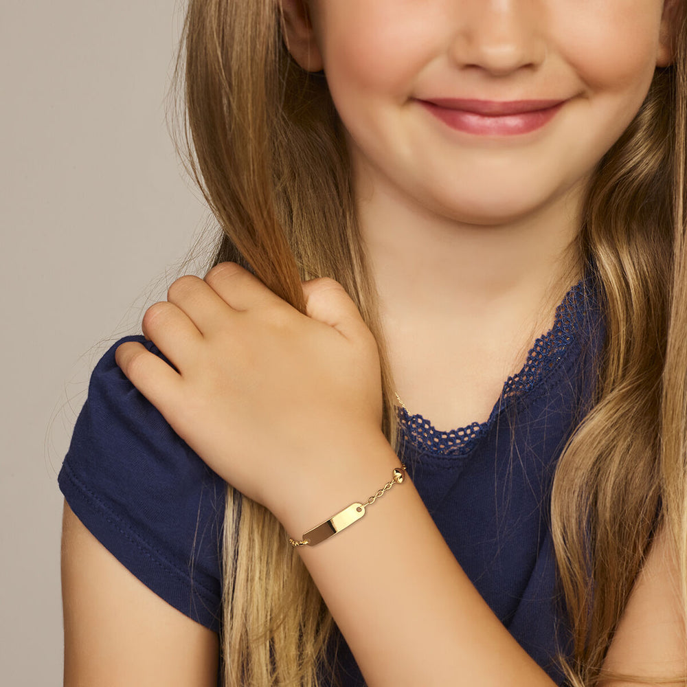 Een jong meisje met lang blond haar en een blauw shirt lacht zachtjes. Een Van der Kooij Collectie 9k grafarmband hartje plaat 4,0 mm 9 Karaat in geelgoudkleur siert haar pols terwijl haar hand op haar schouder rust. De achtergrond is neutraal.