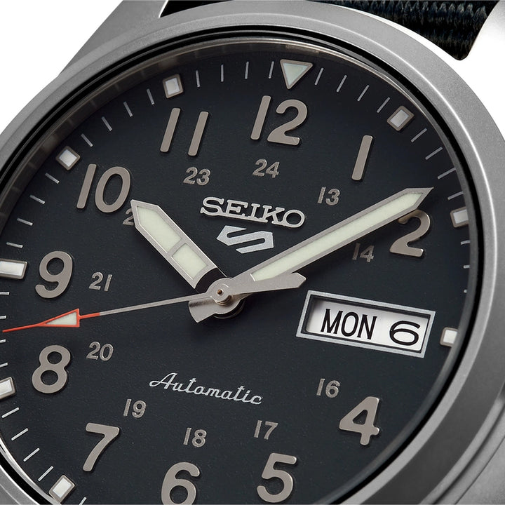 Een close-up van het Seiko 5 Sports herenhorloge SRPG31K1 heeft gloeiende uur- en minutenwijzers, grote Arabische cijfers, een dag-datumweergave met MON 6 en een 24-uursschaal. Het woord Automatic is elegant geplaatst aan de onderkant van dit stijlvolle horloge van Seiko.