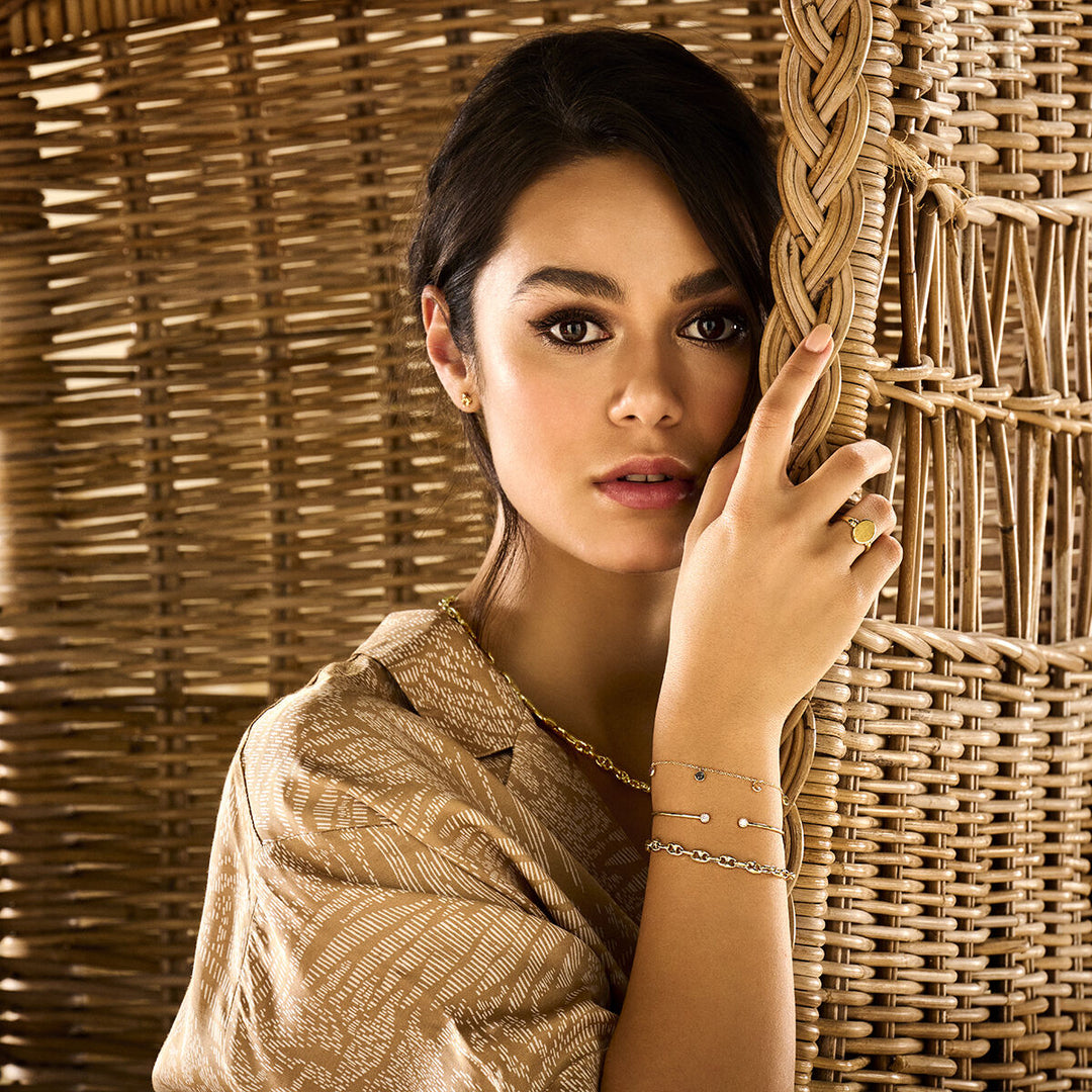 Een donkerharige vrouw poseert bij een rieten scherm, draagt een beige blouse met patroon, gouden sieraden en een Van der Kooij Collectie graveerring massief mat gediamanteerd 14K geelgoud terwijl ze naar de camera kijkt.