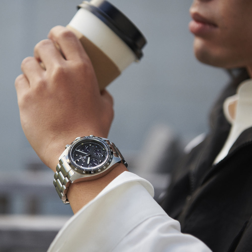 Het individu, getooid met een Seiko Prospex herenhorloge SSC819P1 met een roestvrijstalen kast, houdt een papieren koffiekopje vast met een zwart deksel. Gekleed in een wit overhemd en een donker jasje, is hun gezicht gedeeltelijk zichtbaar tegen de wazige achtergrond.