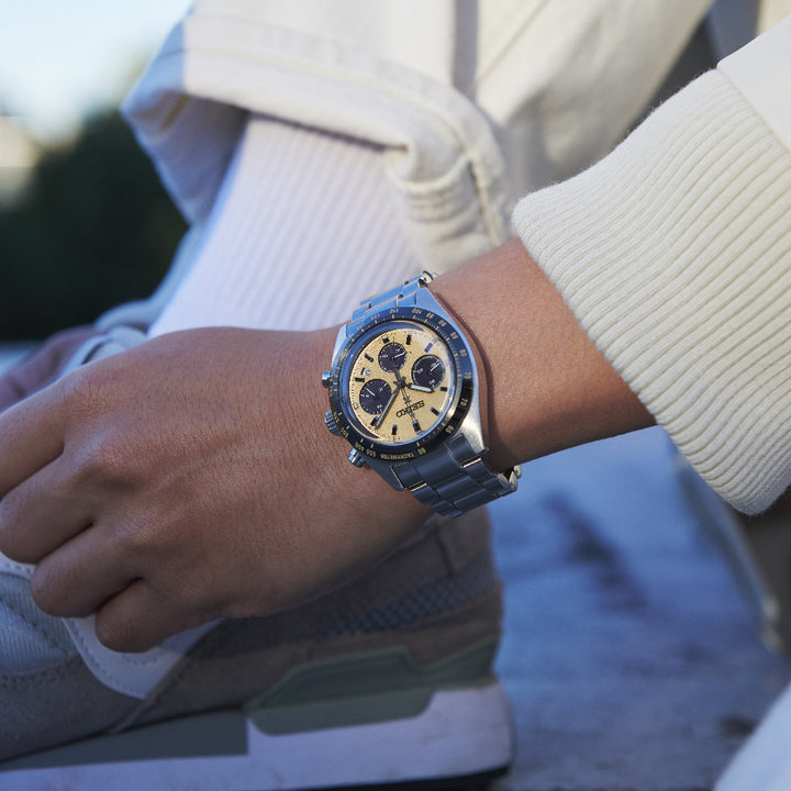 Close-up van een persoon die een Seiko Prospex heren horloge SSC817P1 draagt, met een zilveren afwerking met een beige wijzerplaat en blauwe sub-wijzerplaten. Hun hand rust op hun been, gekleed in een crèmekleurige trui en lichtgekleurde broek, met witte sneakers zichtbaar op de achtergrond.