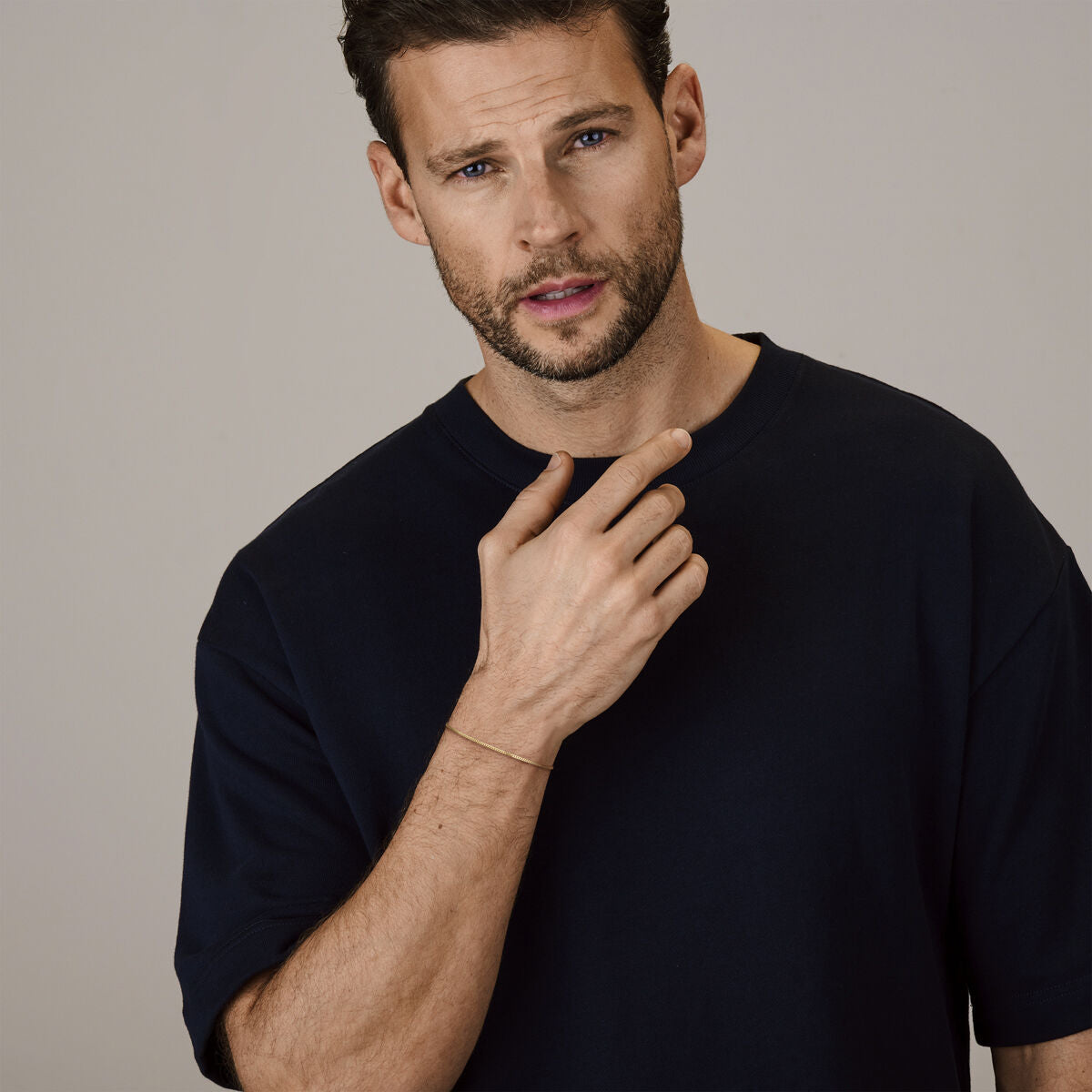 Een man met kort bruin haar en een baard, draagt een donker marineblauw t-shirt, raakt zijn nek aan en toont de Van der Kooij Collectie gourmette 4-zijdes geslepen 1,4 mm 19+2 cm (heren lengte) 14K geelgoud armband tegen een effen achtergrond.