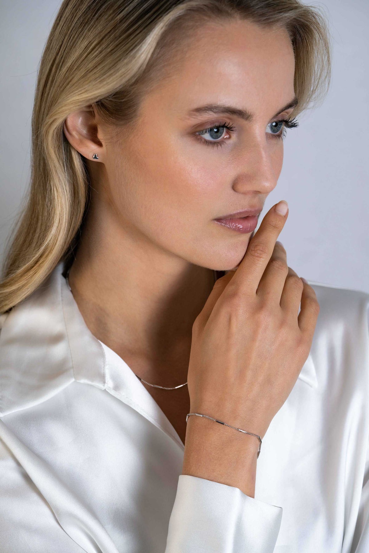 Een vrouw met lang blond haar, gekleed in een witte satijnen blouse, draagt Zinzi Oorbellen Zilver Zio2365 oorbellen, een dunne ketting en een fijne armband terwijl ze nadenkt tegen een neutrale achtergrond.