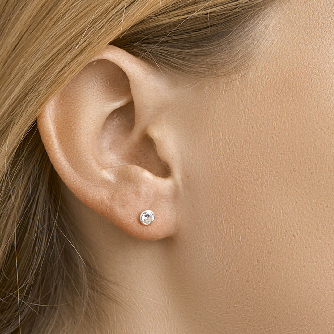 Close-up van een vrouwenoor met steil lichtbruin haar, met oorknoppen zirkonia zilver wit uit de Van der Kooij Collectie - kleine ronde oorstekers met een witte zirkonia steen in een zilveren zetting.
