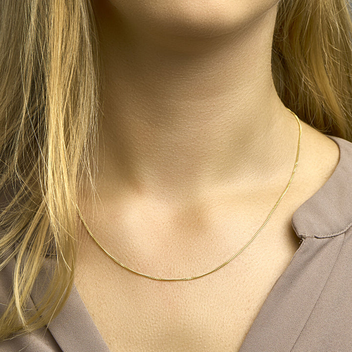 Een close-up van een vrouw in het Van der Kooij Collectie collier gourmette 4-zijden geslepen 1,2 mm 14K geelgoud. Ze heeft lichtblond haar, een taupekleurige blouse met V-hals en staat voor een zacht wazige neutrale achtergrond.