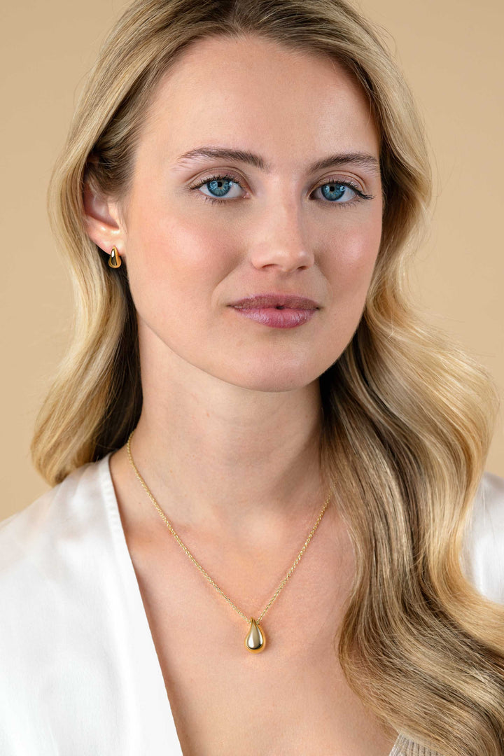 Een vrouw met lang blond haar en blauwe ogen draagt Zinzi Oorbellen Zilver verguld ZIO2636G van Zinzi met een bijpassende gouden traanvormige halsketting. Ze is gekleed in een witte blouse en poseert sierlijk tegen een beige achtergrond.