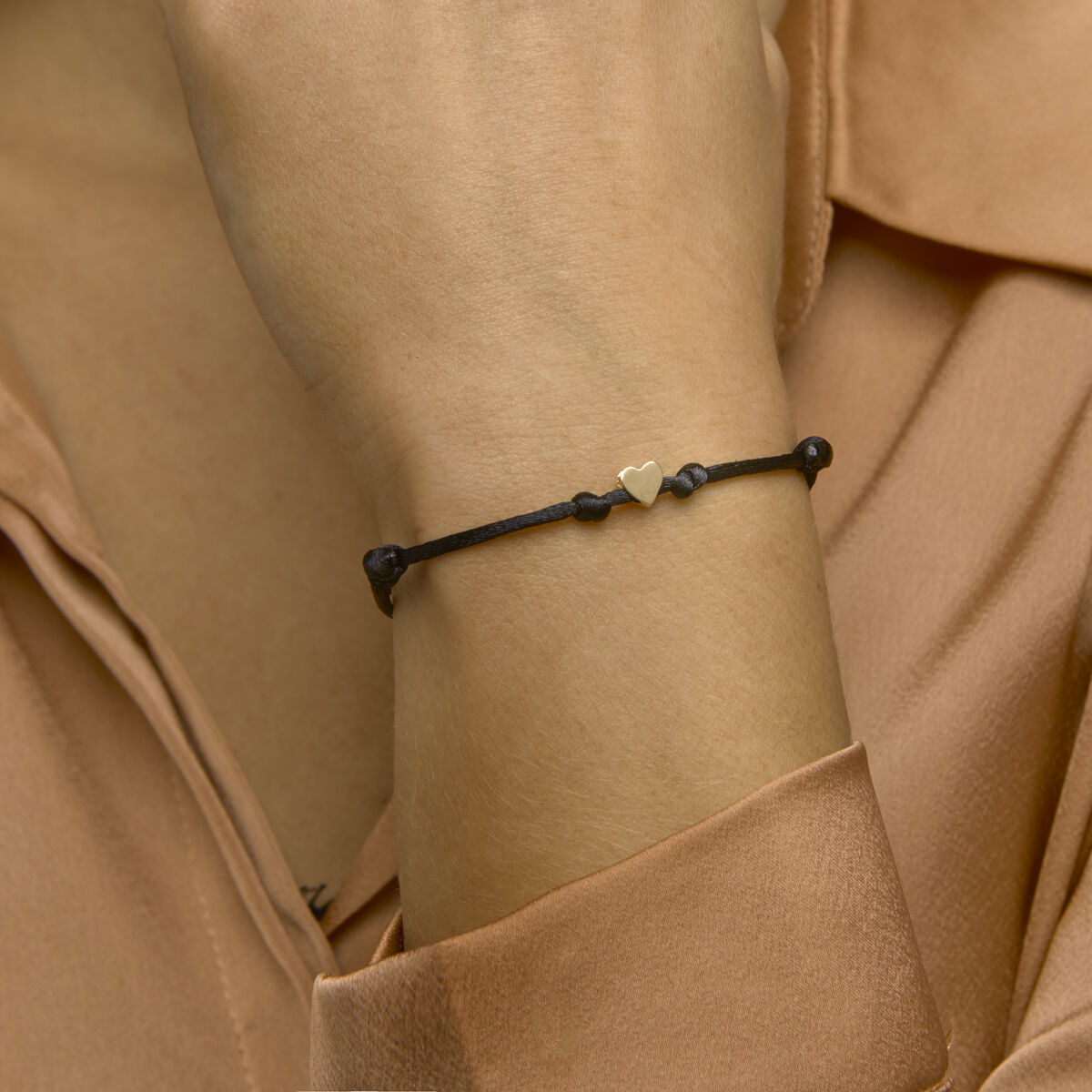 Een persoon in een perzikkleurige blouse draagt de Van der Kooij Collectie armband satijn hart 14K goud met rubber/nylon/leer om zijn pols - een zwart gevlochten bandje met een gouden hartje - terwijl hij zachtjes zijn gezicht aanraakt.
