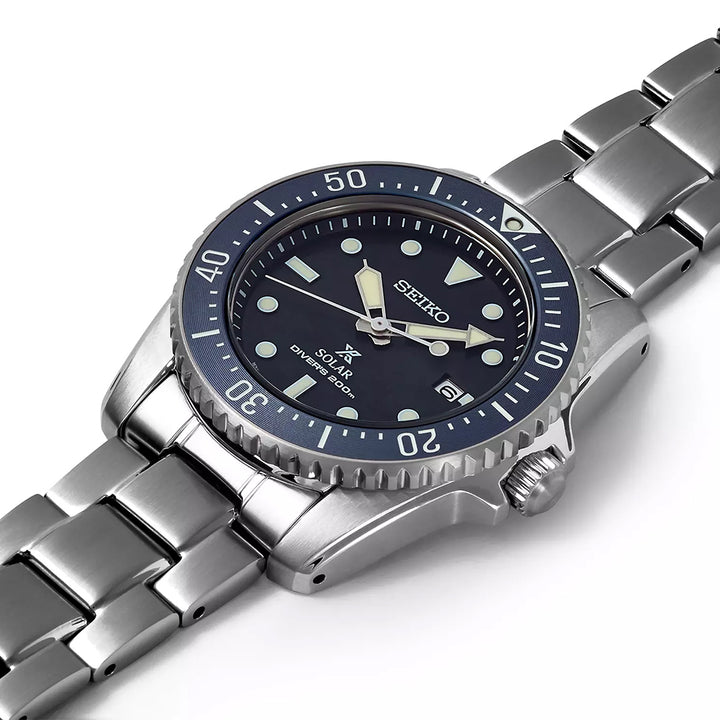 Het Seiko Prospex SNE569P1 Solar Divers 200m horloge heeft een roestvrijstalen kast en een zilveren metalen band. De blauwe wijzerplaat heeft lichtgevende markeringen, wijzers en een datumweergave op 3 uur, terwijl de lunette minutenindicatoren heeft voor nauwkeurige timing.