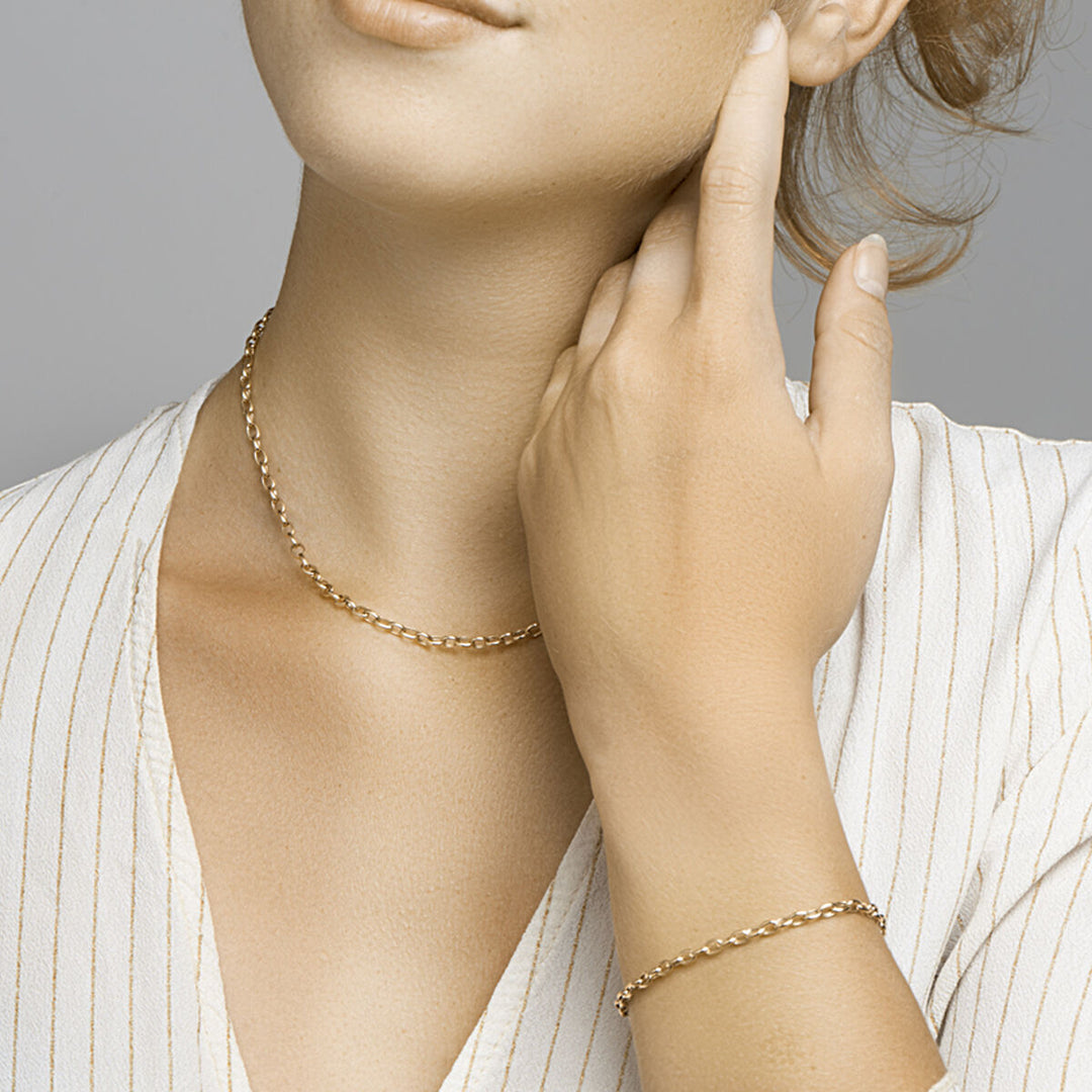 Een vrouw in een witte krijtstreepblouse showt een delicaat gouden kettinkje en de Gouden armband dames anker 14K van Van der Kooij Collectie, haar hand zachtjes in haar nek.