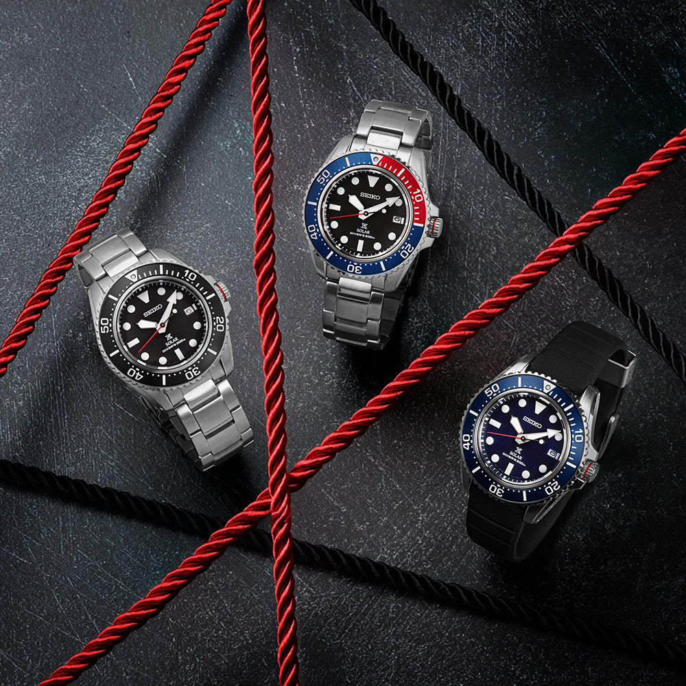 Drie Seiko Prospex heren horloges, model SNE593P1, worden getoond op een donker, gestructureerd oppervlak met rode touwen. Met metalen en rubberen banden, hebben de horloges zwarte wijzerplaten en draaiende bezels in zwart, blauwrood en blauw. De scène benadrukt textuur en contrast.