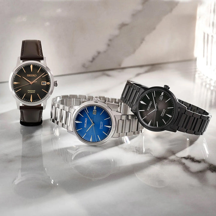 Op een marmeren oppervlak geven drie polshorloges tegelijkertijd dezelfde tijd weer: het Seiko Presage heren horloge SRPJ17J1 met een bruine leren band en zwarte wijzerplaat, een horloge met een zilveren metalen band en een blauwe wijzerplaat en een horloge met een zwarte metalen band en Hardlex mineraalglas, terwijl het licht zachte schaduwen werpt.